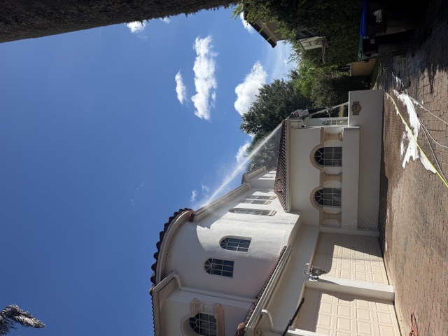 Two-story stucco home being soft washed with a spray arc against a blue sky
